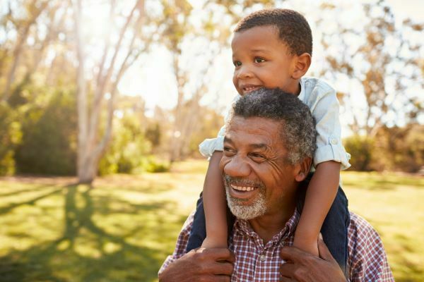 An older black man smiles as his grandson rides on his shoulders